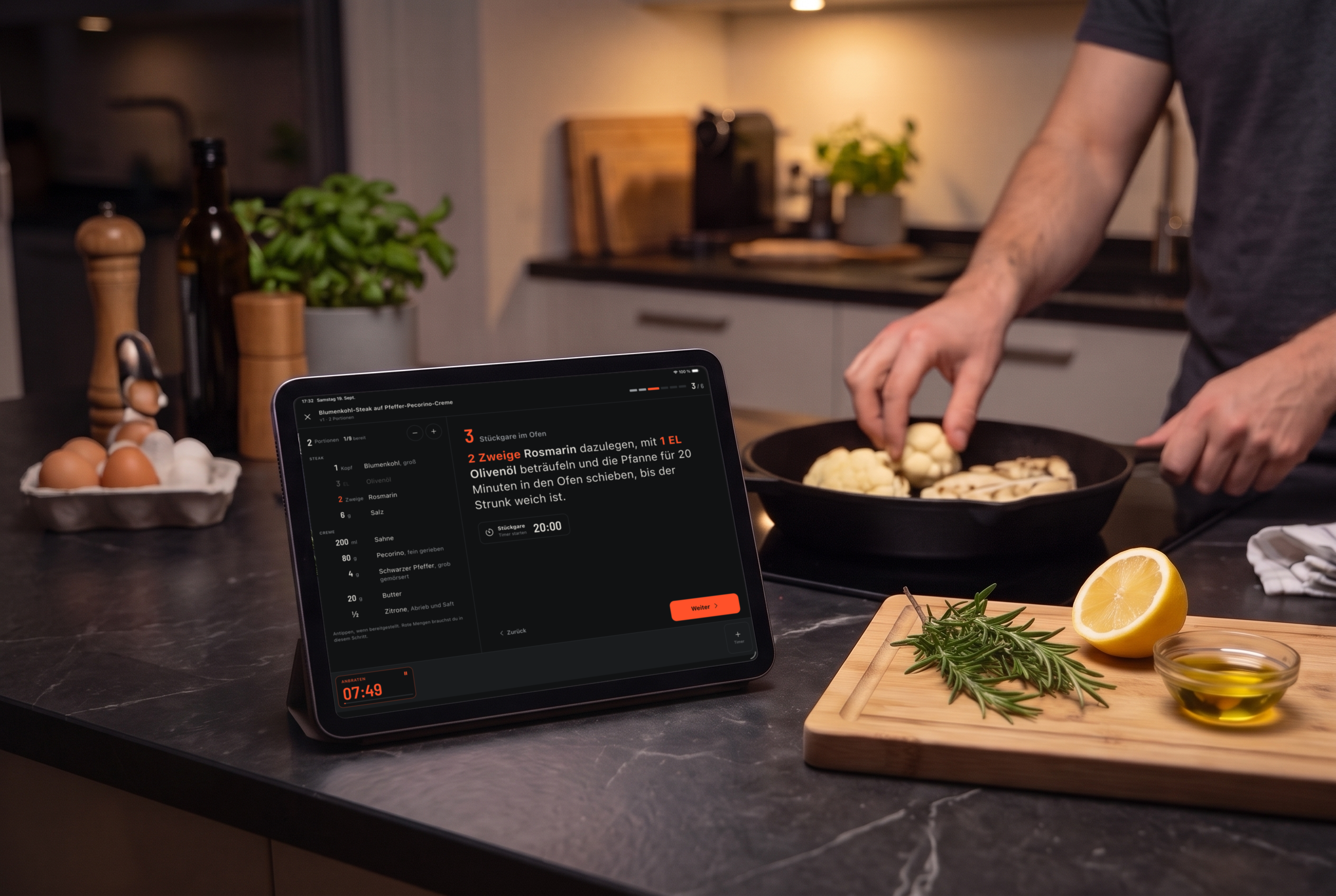 Abends in der Küche: Auf der Arbeitsfläche steht ein Tablet mit dem Kochmodus von Cook & Repeat, dahinter wendet jemand Blumenkohl in einer Gusspfanne.